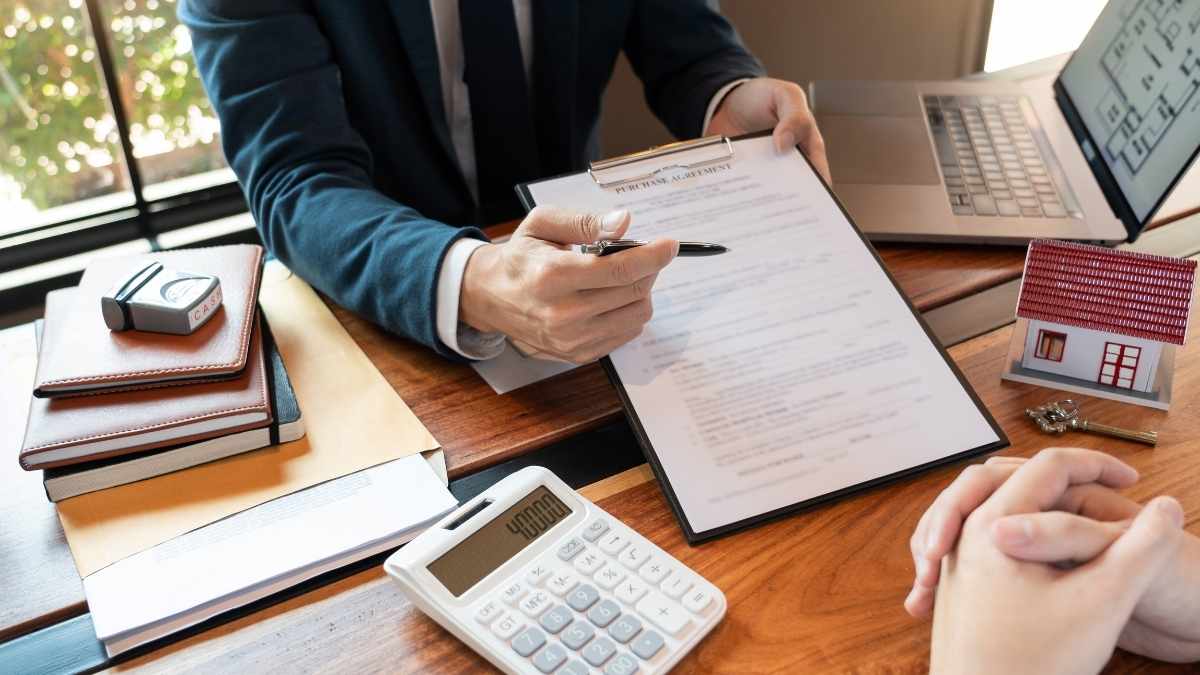 Twee mensen zitten aan het bureau van een advocaat. De handen en armen van de advocaat zijn zichtbaar, van de cliënt zijn enkel de handen zichtbaar. De advocaat heeft een klembord vast en wijst iets aan met een pen. Rechts op tafel van de cliënt staat een klein huisje en ligt een sleutel.