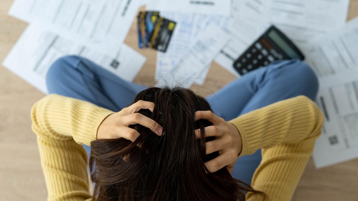 Een vrouw in een blauwe jeansbroek en een gele trui zit in kleermakerszit op de grond met de handen in de haren. Voor haar liggen verschillende rekeningen, twee betaalkaarten en een rekenmachine.