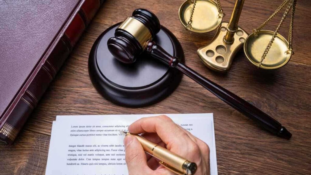 Een houten tafel met een voorzittershamer, boek en weegschaal. Een hand steunt op een bedrukt document. De hand houdt een gouden pen vast.