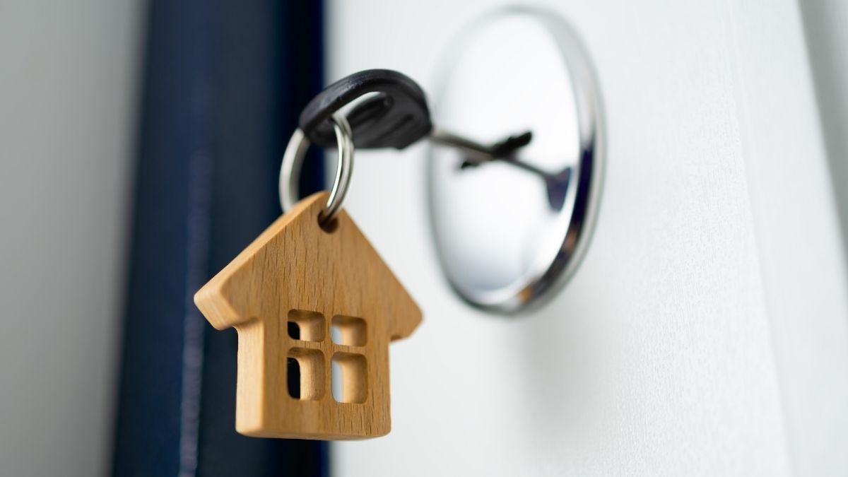 Een sleutelhanger in hout, in de vorm van een klein huisje. De bijhorende sleutel zit in het slot van de witte deur van een huurwoning.