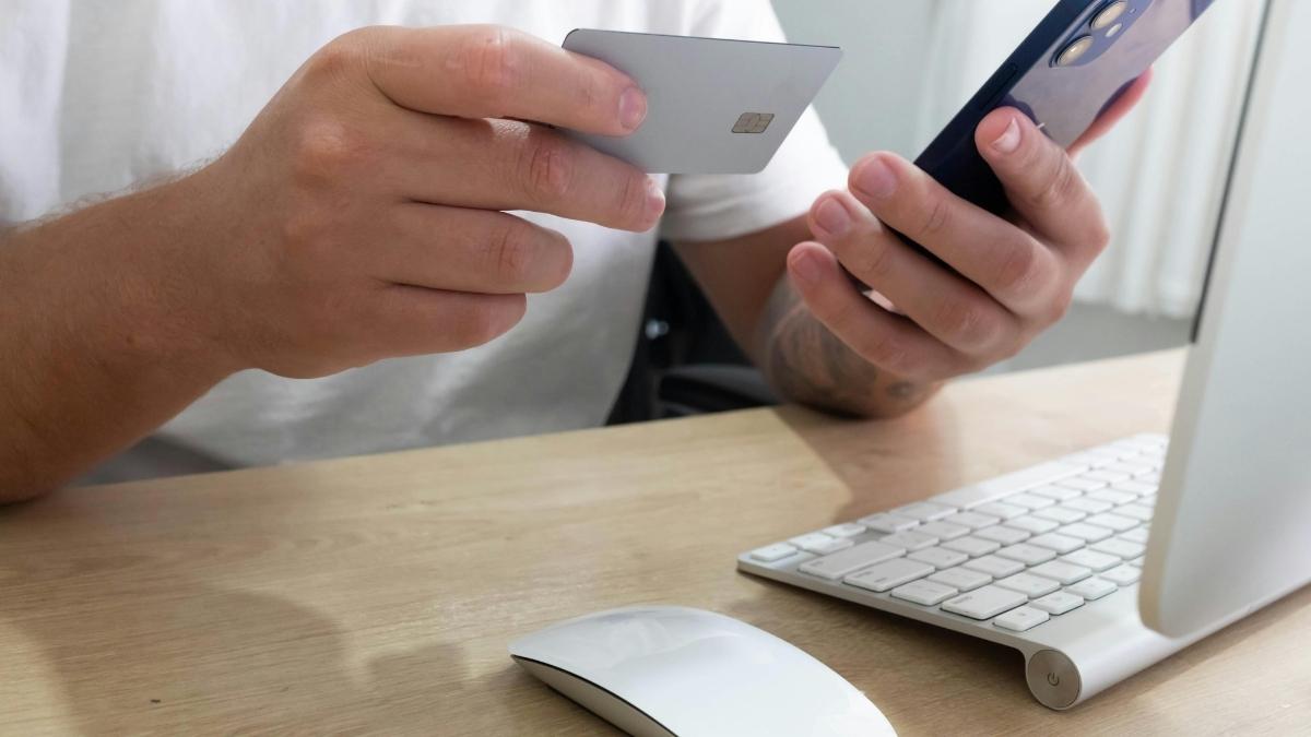 Een man met wit t-shirt zit achter een witte laptop. In zijn linkerhand houdt hij een smartphone vast, met de rechterhand brengt hij zijn witte bankkaart richting de smartphone.