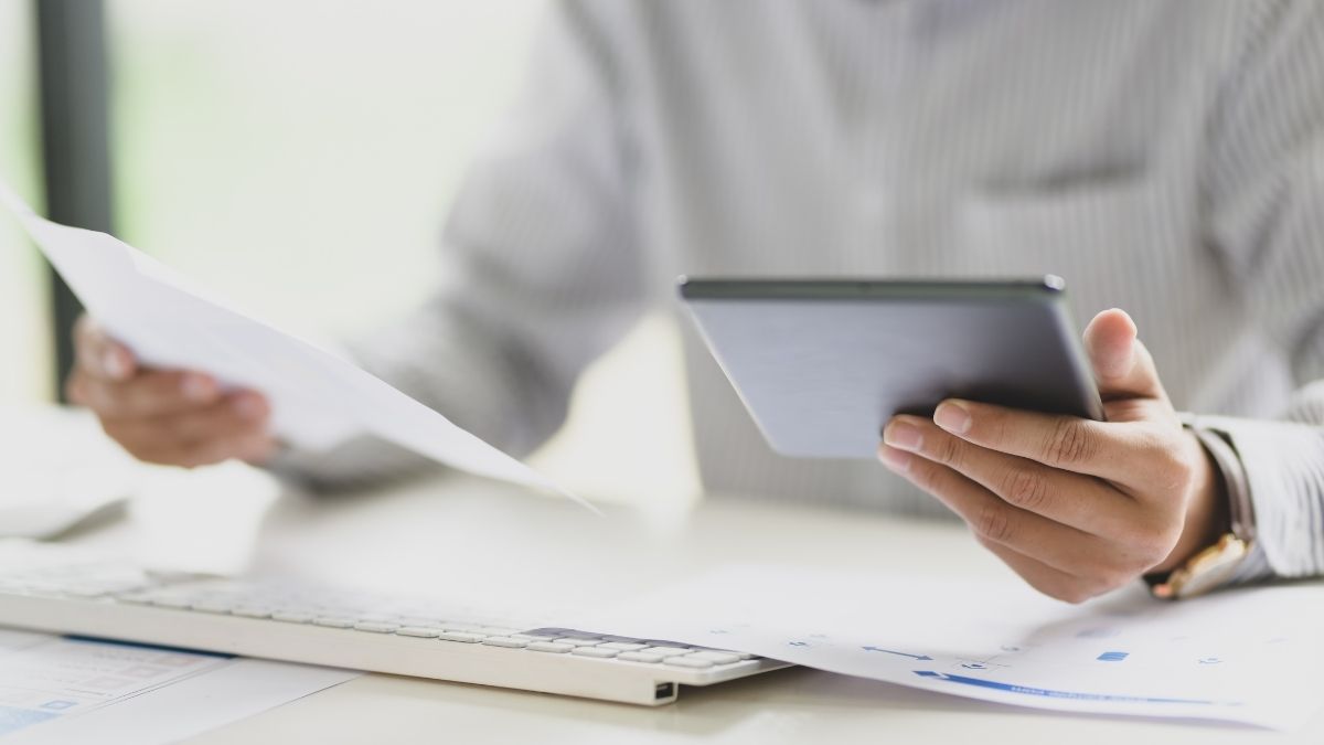 Een persoon in gestreept hemd zit achter een bureau. In de linkerhand houdt hij een tablet vast, in de rechter een blad.