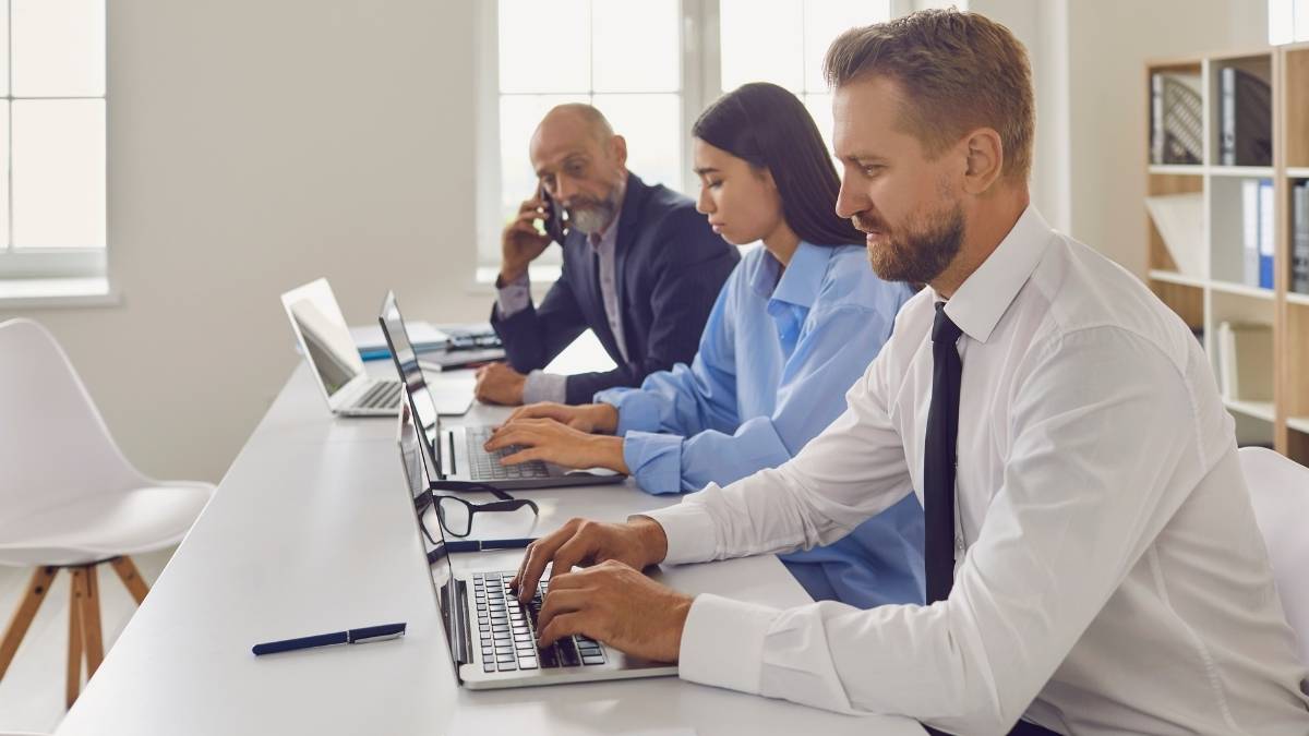 Twee mannen en een vrouw zitten naast elkaar aan tafel. De voorste twee, een man en de vrouw, werken op laptops. De achterste, een oudere man, is aan het telefoneren.