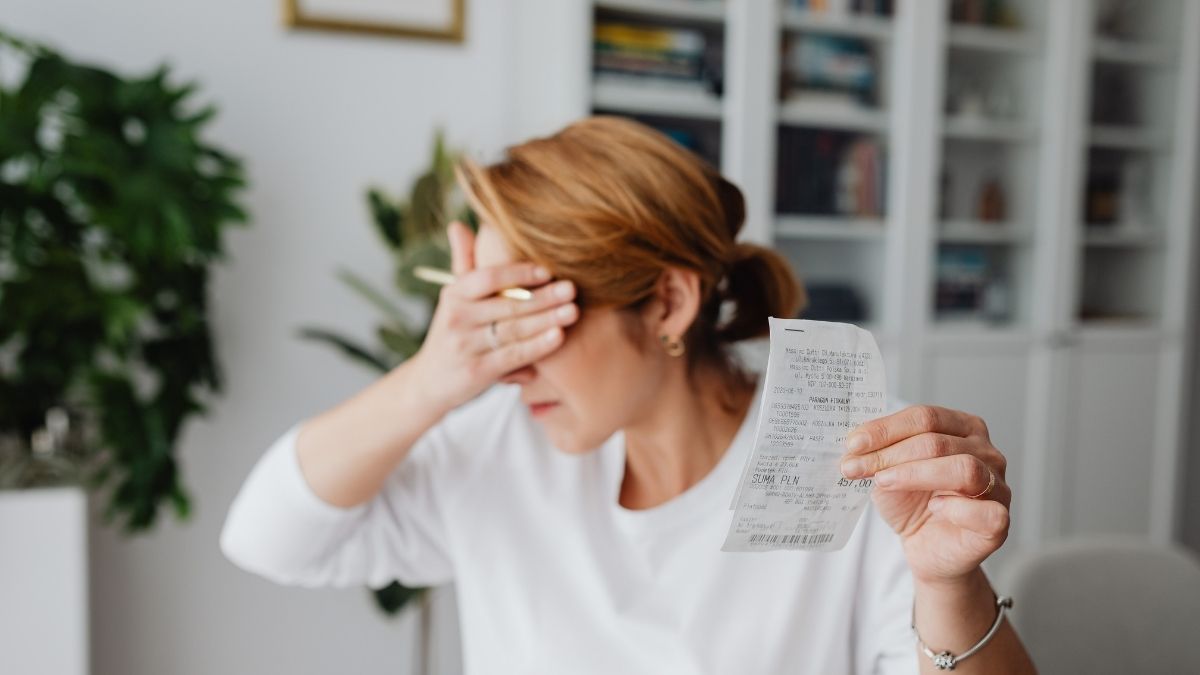 Een vrouw met lichtrood haar en een wit hemd slaat met haar hand tegen haar voorhoofd. Met haar andere hand toont ze een rekening die ze vergat te betalen.